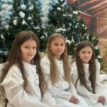 Trzy dziewczynki przebrane za anioły, ubrane w białe, delikatne sukienki, siedzą na tle ośnieżonych choinek i świątecznej dekoracji. Dzieci patrzą w stronę obiektywu z łagodnym, skupionym wyrazem twarzy.