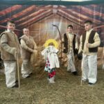 Dzieci z zespołu „Śwarni Lubomiyrzanie” w strojach pasterskich stoją wokół symbolicznej stajenki bożonarodzeniowej. W centrum znajduje się żłóbek z Dzieciątkiem, a całość dopełnia świąteczna scenografia z choinką, sianem i dekoracyjnym tłem imitującym wnętrze szop