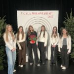 Grupa uczennic oraz opiekunka stoją na scenie podczas V Gali Wolontariatu 2025; w tle dekoracja z logo wydarzenia i choinki.