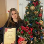 Uczennica trzyma dyplom „Wolontariusz Roku 2025” oraz kwiaty, stojąc obok udekorowanej choinki świątecznej.