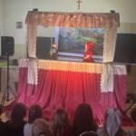 Dzieci siedzą na krzesłach i oglądają przedstawienie kukiełkowe na szkolnej scenie z dekoracją leśną.