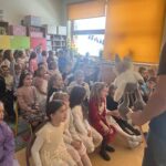 Dzieci siedzą na krzesłach i oglądają występ z pacynką prowadzony przez nauczyciela w szkolnej sali.