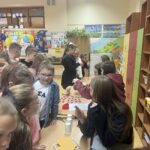 Dzieci stoją przy stoliku z kartami i kubkami, biorąc udział w andrzejkowych zabawach wróżbiarskich w szkolnej klasie.