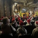 Dzieci siedzą w sali podziemnej kopalni, zwrócone w stronę Świętego Mikołaja stojącego przy dekoracjach świątecznych