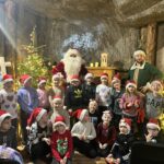 Grupa dzieci w czapkach mikołajowych pozuje z postacią Świętego Mikołaja i pomocnikiem w świątecznie ozdobionej komorze solnej.