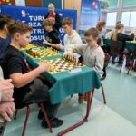 Uczniowie grają w szachy przy stolikach w sali szkolnej, skupieni na partiach rozgrywanych podczas turnieju.