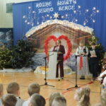 Uroczystość szkolna na sali gimnastycznej. Dyrektorka przemawia przy mównicy na tle świątecznej dekoracji z szopką, choinkami i napisem „Różne światy jedno serce”. Obok stoją dzieci w strojach ludowych i galowych, część z instrumentami, a przed sceną zgromadzona jest publiczność uczniów.”