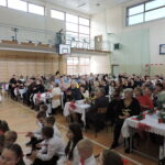 Publiczność zgromadzona w sali gimnastycznej podczas uroczystości szkolnej. Przy długich stołach siedzą rodzice, dziadkowie i zaproszeni goście, a na pierwszym planie widoczne są dzieci w galowych strojach. Uczestnicy wydarzenia biją brawo, oglądając występ na scenie.