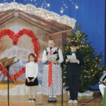 Dzieci występują na szkolnej scenie podczas uroczystości bożonarodzeniowej. Uczennica w stroju ludowym i uczeń w stroju galowym czytają teksty do mikrofonów, obok stoi młodsza dziewczynka. W tle widoczna jest świąteczna dekoracja z szopką, choinką i sercem z czerwonych świateł.