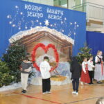 Dzieci na szkolnej scenie przygotowują się do występu bożonarodzeniowego. Przed świąteczną dekoracją z szopką, choinkami i napisem „Różne świąty jedno serce” uczniowie ustawiają się w strojach galowych i ludowych, część z instrumentami muzycznymi.