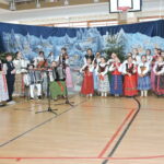 Grupa uczniów występuje na szkolnej scenie podczas uroczystości bożonarodzeniowej. Dzieci w strojach ludowych śpiewają i grają na instrumentach, m.in. skrzypcach i akordeonach, na tle zimowej dekoracji z choinkami i świąteczną scenerią.