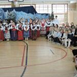 Uczniowie zgromadzeni na sali gimnastycznej podczas szkolnej uroczystości bożonarodzeniowej. Na scenie stoją dzieci w strojach ludowych, część z instrumentami, a naprzeciw nich siedzą młodsi uczniowie oraz opiekunowie, oglądający występ artystyczny.