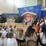 Występ bożonarodzeniowy w sali gimnastycznej. Dzieci w strojach ludowych i galowych prezentują program artystyczny na tle świątecznej dekoracji z szopką i choinkami, a przed sceną zgromadzeni są młodsi uczniowie oraz opiekunowie oglądający występ.