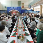 Widok sali gimnastycznej podczas szkolnej uroczystości bożonarodzeniowej. Przy długich, świątecznie udekorowanych stołach siedzą licznie zgromadzeni goście, a w tle na scenie uczniowie prezentują program artystyczny na tle szopki i świątecznych dekoracji.