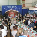 Licznie zgromadzona publiczność w sali gimnastycznej podczas szkolnej uroczystości bożonarodzeniowej. Przy długich, świątecznie udekorowanych stołach siedzą goście, a na scenie uczniowie w strojach ludowych i galowych występują na tle dekoracji z szopką, choinkami i napisem „Różne światy, jedno serce”.