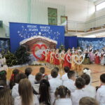 Występ bożonarodzeniowy w sali gimnastycznej. Grupa uczennic w złoto-czerwonych strojach tanecznych prezentuje układ z dużymi rekwizytami w kształcie serc, a w tle pozostali uczniowie w strojach ludowych śpiewają i grają na instrumentach przy świątecznej dekoracji z szopką i choinkami.