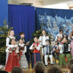Uczniowie występują na szkolnej scenie podczas uroczystości bożonarodzeniowej. Dzieci w tradycyjnych strojach ludowych grają na instrumentach, m.in. skrzypcach, wiolonczeli i akordeonach, na tle zimowej dekoracji z choinkami i świąteczną scenerią.