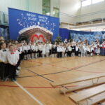 Wspólny występ uczniów w sali gimnastycznej podczas szkolnej uroczystości bożonarodzeniowej. Dzieci w galowych strojach stoją w długim rzędzie i śpiewają kolędy na tle świątecznej dekoracji z szopką, choinkami i napisem „Różne światy, jedno serce”.