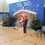 Dyrektorka szkoły przemawia do zgromadzonych podczas uroczystości bożonarodzeniowej w sali gimnastycznej. Stoi z mikrofonem na tle świątecznej dekoracji z szopką, choinkami i napisem „Różne światy, jedno serce”.