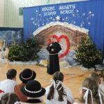 Duchowny przemawia do zgromadzonych podczas szkolnej uroczystości bożonarodzeniowej w sali gimnastycznej. Stoi z mikrofonem na tle świątecznej dekoracji z szopką, choinkami i napisem „Różne świąty, jedno serce”, a przed nim siedzą uczniowie w strojach galowych i ludowych.