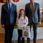 Uczennica szkoły podstawowej stoi pomiędzy dwoma dorosłymi mężczyznami na tle baneru Powiatu Limanowskiego. Dziewczynka trzyma dyplom oraz torbę z logo powiatu. Wszyscy pozują do pamiątkowego zdjęcia po wręczeniu nagrody.