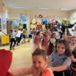 Uczniowie klas I–III w przebraniach karnawałowych biorą udział we wspólnej zabawie tanecznej w sali szkolnej, trzymając się za ramiona i poruszając w rytm muzyki.
