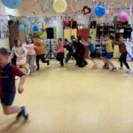 Uczniowie klas I–III w przebraniach karnawałowych biorą udział we wspólnej zabawie tanecznej w sali szkolnej, trzymając się za ramiona i poruszając w rytm muzyki.