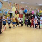 Uczniowie klas I–III w przebraniach karnawałowych biorą udział we wspólnej zabawie tanecznej w sali szkolnej, trzymając się za ramiona i poruszając w rytm muzyki.
