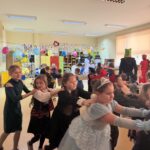 Uczniowie klas I–III w przebraniach karnawałowych biorą udział we wspólnej zabawie tanecznej w sali szkolnej, trzymając się za ramiona i poruszając w rytm muzyki.