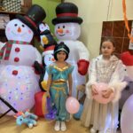 Dwie uczniów klas I–III w strojach karnawałowych stoi przed dekoracją z dmuchanymi postaciami i trzyma kolorowe balony podczas balu karnawałowego.