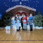 Uczniowie z dyplomami stoją razem z nauczycielkami w sali gimnastycznej po szkolnej uroczystości bożonarodzeniowej. Zdjęcie wykonane na tle świątecznej dekoracji z szopką, choinkami, sercem z czerwonych ozdób oraz napisem „Różne światy, jedno serce”.