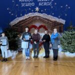 Grupowe zdjęcie uczniów i nauczycielek w sali gimnastycznej po szkolnej uroczystości bożonarodzeniowej. Uczniowie trzymają dyplomy i torby z upominkami, stojąc na tle świątecznej dekoracji z szopką, choinkami i napisem „Różne światy, jedno serce”.