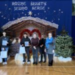 Grupowe zdjęcie uczniów i nauczycieli na tle świątecznej dekoracji w sali gimnastycznej. Dwóch uczniów trzyma dyplomy i torby z upominkami, a obok stoją nauczycielki. W tle widoczna jest szopka, choinki oraz napis „Różne światy, jedno serce”.
