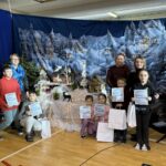 Grupowe zdjęcie uczniów i nauczycieli po szkolnej uroczystości bożonarodzeniowej. Dzieci trzymają dyplomy i torby z upominkami, stojąc i klęcząc przed świąteczną dekoracją z szopką, choinkami i zimową scenerią.