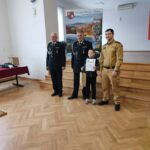 Uczeń odbiera dyplom i nagrodę podczas etapu gminnego Ogólnopolskiego Turnieju Wiedzy Pożarniczej. Obok stoją strażacy w galowych mundurach oraz przedstawiciel straży w umundurowaniu służbowym.