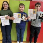 Troje uczniów prezentuje dyplomy oraz nagrody zdobyte w części czytelniczej konkursu „Góralskie Cytanie”. Zdjęcie wykonano w szkolnej sali na tle kolorowych szafek.