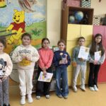 Uczennice z młodszych klas stoją w szkolnej sali na tle kolorowej ilustracji z postaciami z bajki. Niektóre z nich trzymają dyplomy i nagrody zdobyte w konkursie „Góralskie Cytanie”.