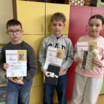 Troje uczniów prezentuje dyplomy i nagrody zdobyte w części plastycznej konkursu „Góralskie Cytanie”. Uczniowie stoją przed kolorowymi szafkami w szkolnej sali.