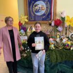 Laureatka konkursu „Jajko Wielkanocne w Objęciach Wiosny” podczas wręczenia dyplomu i nagrody. W tle widoczna jest wystawa konkursowych prac – kolorowe pisanki, wiosenne kwiaty i wielkanocne dekoracje przygotowane przez uczniów.