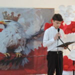 Chłopiec w galowym stroju szkolnym recytuje tekst przy mikrofonie na tle dużego obrazu białego orla w koronie symbolizującego Polskę