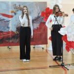 Dwie uczennice wykonują występ muzyczny – jedna śpiewa, a druga gra na skrzypcach podczas szkolnej akademii patriotycznej