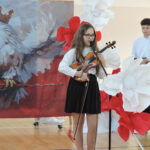 Dziewczynka trzyma skrzypce i śpiewa do mikrofonu, uczestnicząc w szkolnej akademii o tematyce patriotycznej