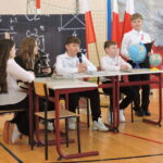 Grupa uczniów siedzi przy szkolnych ławkach i prowadzi prezentację edukacyjną z wykorzystaniem globusa, mikroskopu i pomocy naukowych