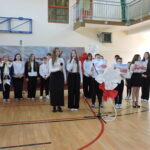 Grupa uczniów wspólnie występuje na zakończenie szkolnej akademii patriotycznej w udekorowanej sali gimnastycznej