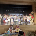 Uczennice i uczniowie w regionalnych strojach stoją na scenie obok dekoracji kwiatowej, trzymając nagrody konkursowe.