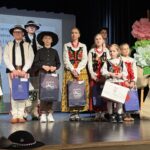 Grupa uczestników konkursu w strojach ludowych na scenie, poniżej widoczne dekoracje z lalkami i publikacjami regionalnym