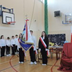 Poczet sztandarowy wraz z uczniami stoi w sali gimnastycznej udekorowanej na szkolną akademię o charakterze patriotycznym