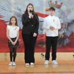 Troje uczniów stoi na środku sali gimnastycznej i recytuje wiersze o tematyce patriotycznej podczas uroczystości szkolnej