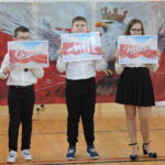 Troje uczniów trzyma plansze z napisami „Ojczyzna”, „Naród” i „Wolność” na tle dekoracji z orłem i barwami narodowymi