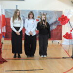 Trzy uczennice stoją obok siebie, a jedna z nich przemawia do mikrofonu, recytując wiersz o tematyce patriotycznej, w tle znajduje się dekoracja przedstawiająca białego orła w koronie